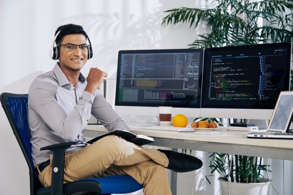 Portrait of smiling confident Indian coder sitting at his office desk with programming code on computer screens