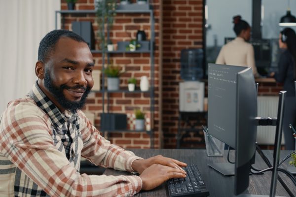 Database programer writing code and smiling while coworker walks by holding clipboard in it agency. Relaxed app developer typing on keyboard with colleagueas computing big data.