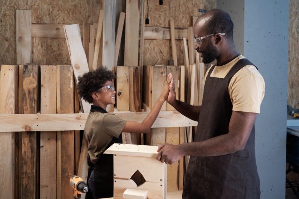 Side view portrait of father and son high five while building wooden birdhouse in workshop together